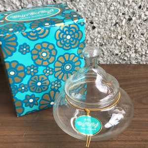 Puede incluir: Un tarro de vidrio transparente con tapa y una cuerda dorada atada alrededor del cuello. El tarro contiene cápsulas de baño y tiene una etiqueta que dice "Wyott Skin's Soft Bath Capsules". El tarro está sentado sobre una superficie de madera junto a una caja azul y dorada con un patrón floral.