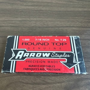 Peut inclure: Une boîte rectangulaire d'agrafes Arrow avec un design rouge et noir. La boîte indique "1 000 7/16 INCH ROUND TOP GENUINE ARROW Staples PRECISION MADE". La boîte est sur une surface en bois.