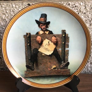 May include: A decorative plate depicting a historical scene of a man in stocks, holding a sign that reads "pe Blutter". The plate has a gold rim with a rope design and a light blue background.