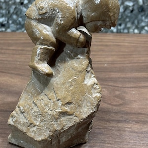 Può includere: Una scultura color marrone chiaro di una figura che scala una formazione rocciosa. La figura è piegata, indossa stivali e un cappello. La roccia ha una superficie ruvida e strutturata e la scultura è posta su una superficie di legno.