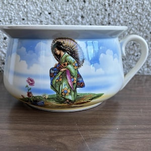 Op de afbeelding: Een witte keramische chamberpot met een blauw en wit bloemenmotief. De pot heeft een vrouw in een groene en roze kimono die een paraplu vasthoudt.