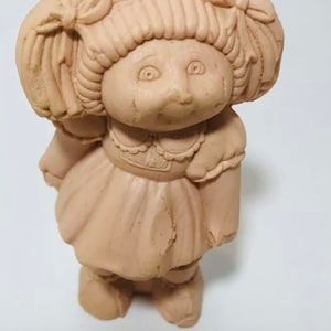 May include: A tan ceramic figurine of a girl with pigtails wearing a dress and a hat.