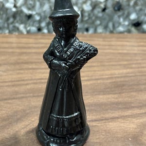 May include: A small, black figurine of a woman in a long dress and hat, holding a bouquet of flowers. The figurine has a glossy finish and is standing on a wooden surface. The figure is a decorative item.