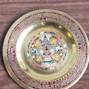 May include: A small, round, gold-colored metal plate with a colorful design of the Aztec calendar. The plate has a red border with black and white writing around the edge. The center of the plate features a colorful design of the Aztec calendar with a red, black, and white face in the center. The plate is inscribed with the word "MEXICO" in red letters.