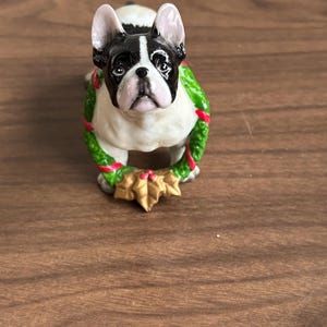 Peut inclure: Figurine en céramique d'un bouledogue français noir et blanc, orné d'une couronne verte et rouge. La couronne est ornée de feuilles de houx dorées. Cette figurine de chien est un objet décoratif.