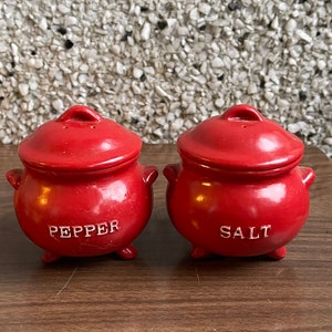 May include: Red ceramic salt and pepper shakers shaped like small pots with lids. The shakers have white lettering that reads "SALT" and "PEPPER".