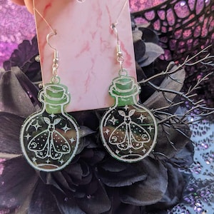 May include: A pair of green glass bottle earrings featuring a white moth design and glitter accents. The earrings are displayed on a white card with a pink marble pattern.