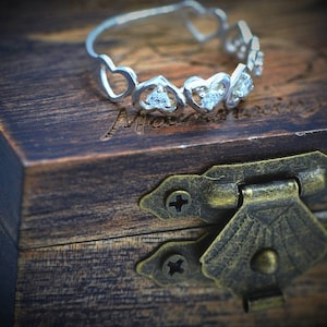 Può includere: Un anello d'argento con un design a forma di cuore e piccole pietre trasparenti. L'anello è su una scatola di legno con una fibbia in ottone.