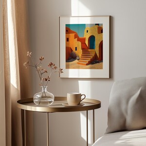 May include: A framed art print depicts a stylized Mediterranean village scene. The artwork features buildings in shades of yellow and orange, a blue door, and a staircase. The print is displayed on a wall above a gold side table with a vase and a cup.