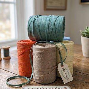 Puede incluir: Cuatro bobinas cilíndricas de cordel de papel en colores verde azulado, naranja, beige y amarillo pálido. Una etiqueta de papel blanco unida a la bobina amarilla dice ECO-TWINE. Un letrero de madera muestra el texto yarnofanatolia HOBBY & CO.