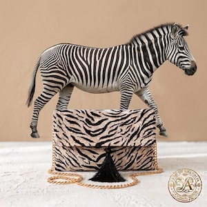 May include: A rectangular clutch bag featuring a beige and black zebra stripe pattern, a black tassel centered on the front flap, and a gold-tone metal chain shoulder strap. A zebra figure stands directly behind the clutch bag.