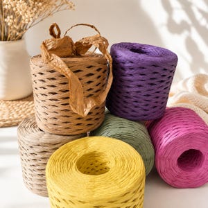 May include: A collection of six cylindrical spools of raffia ribbon in various colors including tan, purple, sage green, bright yellow, and magenta. The spools feature a textured, woven paper construction with a loose strand of tan raffia on top.