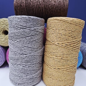 Può includere: Bobine di spago in vari colori, tra cui grigio, beige e marrone, sono disposte su uno sfondo blu. Lo spago è ben avvolto, mostrando una superficie strutturata. L'immagine mette in evidenza le diverse tonalità e trame dello spago.