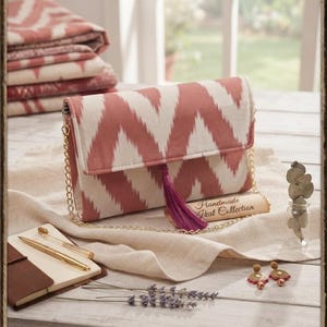 May include: A rectangular clutch bag featuring a coral and cream chevron ikat pattern, a gold chain strap, and a deep purple tassel. A small tag attached to the bag displays the text Handmade Ikat Collection, and a label at the bottom reads IKAT CRAFT COLLECTION - 2023.
