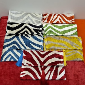 Puede incluir: Una colección de bolsos de mano rectangulares con estampado de cebra. Los bolsos están disponibles en varios colores, como gris, negro, azul, naranja, verde, amarillo y rojo. Algunos bolsos tienen una borla. Los bolsos se muestran sobre una superficie roja.