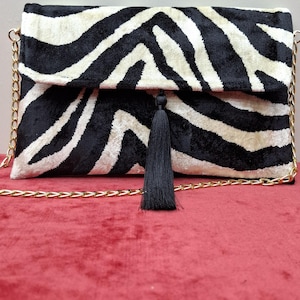 Puede incluir: Un bolso de mano rectangular con un patr&oacute;n de rayas de cebra en blanco y negro con textura aterciopelada. El bolso incluye una correa de cadena de metal dorado y una borla de tela negra centrada en el cierre de solapa frontal.