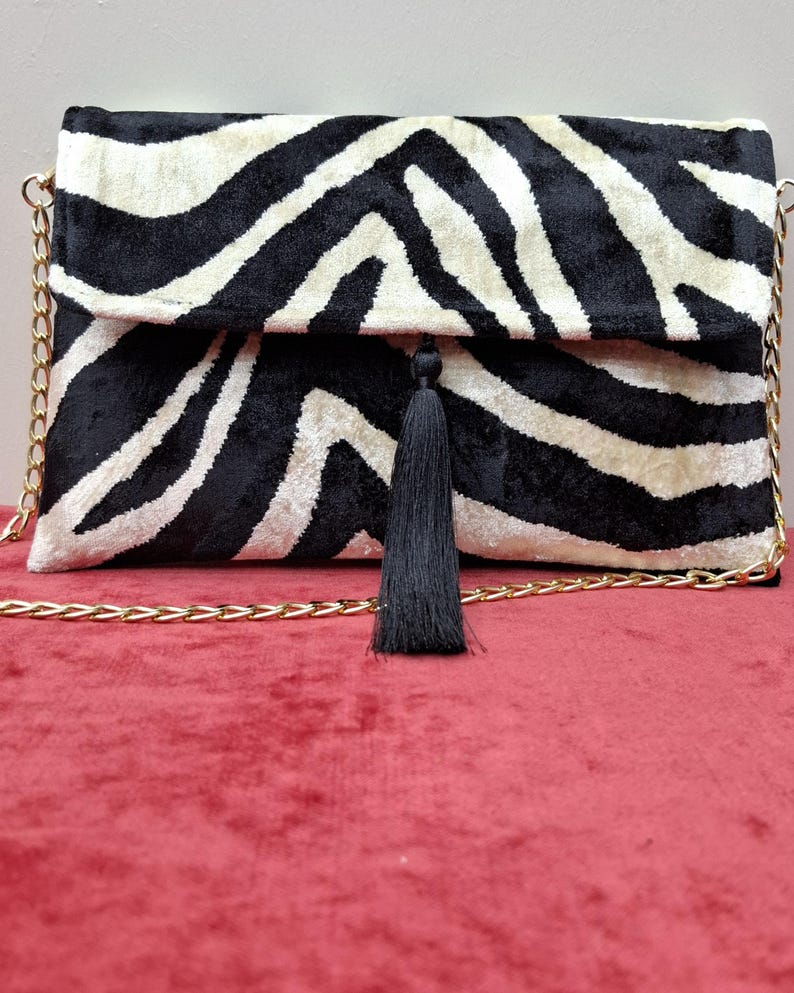 Puede incluir: Un bolso de mano rectangular con un patr&oacute;n de rayas de cebra en blanco y negro con textura aterciopelada. El bolso incluye una correa de cadena de metal dorado y una borla de tela negra centrada en el cierre de solapa frontal.
