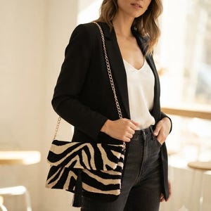 Puede incluir: Un bolso de mano rectangular con un patr&oacute;n de rayas de cebra en negro y beige, una correa de cadena de metal dorado y una borla negra que cuelga de la solapa frontal. El bolso est&aacute; confeccionado con un material suave y texturizado.