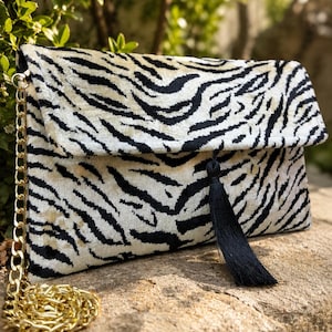 Puede incluir: Un bolso de mano rectangular con un patrón de rayas de cebra en negro y crema sobre una tela similar al terciopelo. El bolso incluye una correa de cadena de metal dorado y una borla decorativa negra larga unida al cierre de solapa frontal.