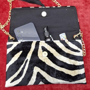 Puede incluir: Un bolso de mano rectangular con estampado de cebra en negro y crema. El bolso tiene una correa de cadena dorada, una borla negra y un cierre magn&eacute;tico. En su interior contiene un tel&eacute;fono inteligente, un peque&ntilde;o pulverizador y un tubo.
