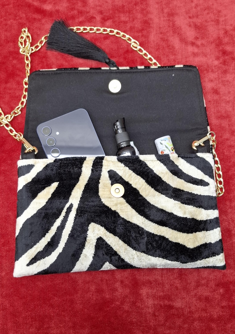 Puede incluir: Un bolso de mano rectangular con estampado de cebra en negro y crema. El bolso tiene una correa de cadena dorada, una borla negra y un cierre magn&eacute;tico. En su interior contiene un tel&eacute;fono inteligente, un peque&ntilde;o pulverizador y un tubo.