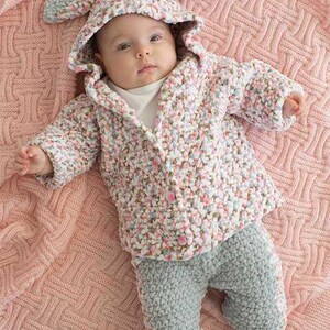 May include: A baby in a handmade bunny-eared hooded jacket and grey trousers, lying on a pink knitted blanket. The jacket has a speckled pattern of pink, white, and blue. The outfit is buttoned up.
