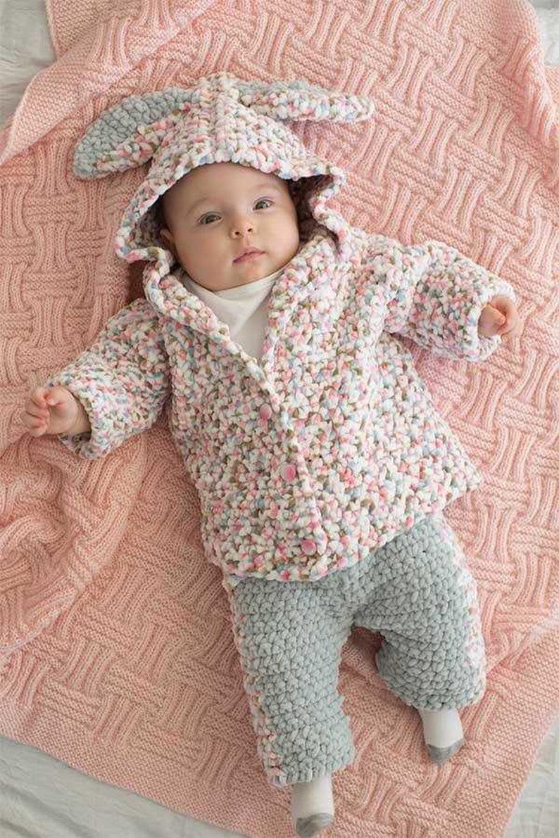 May include: A baby in a handmade bunny-eared hooded jacket and grey trousers, lying on a pink knitted blanket. The jacket has a speckled pattern of pink, white, and blue. The outfit is buttoned up.