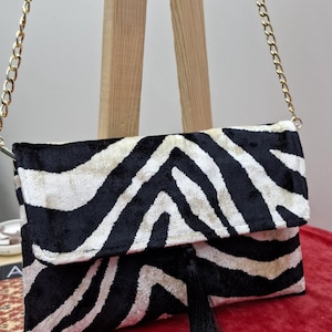 Puede incluir: Un bolso de mano rectangular con un patr&oacute;n de rayas de cebra en blanco y negro con textura aterciopelada. El bolso incluye una correa de cadena de metal dorado y una borla negra larga que cuelga del centro del cierre de solapa frontal.