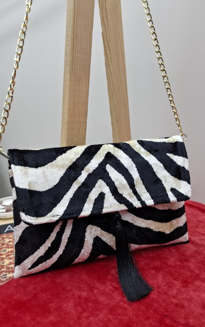 Puede incluir: Un bolso de mano rectangular con un patr&oacute;n de rayas de cebra en blanco y negro con textura aterciopelada. El bolso incluye una correa de cadena de metal dorado y una borla negra larga que cuelga del centro del cierre de solapa frontal.