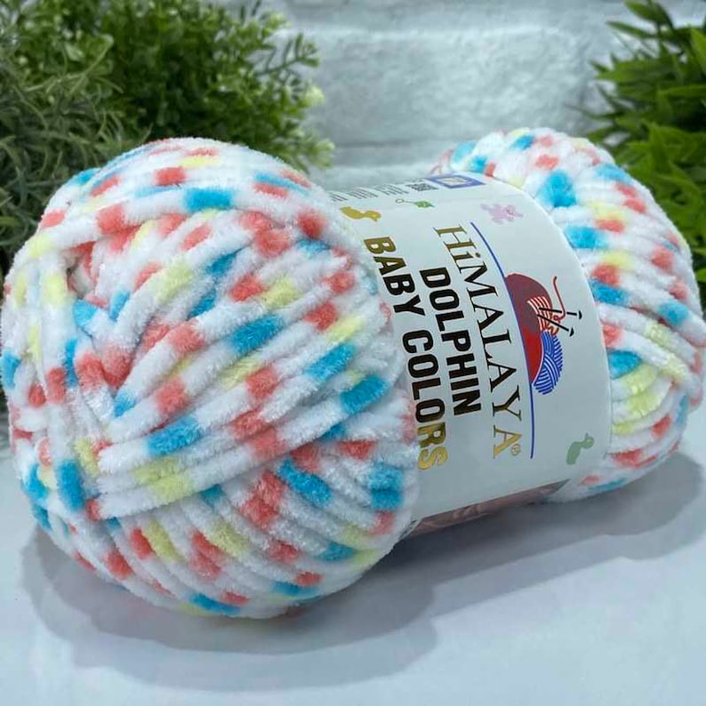 May include: A ball of Himalaya Dolphin Baby Colors yarn. The yarn is white with flecks of blue, yellow, and coral. The label reads "Himalaya Dolphin Baby Colors". The yarn is soft and fluffy, suitable for baby projects.