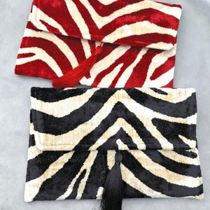 May include: Two rectangular clutch bags with a zebra stripe pattern. One is red and cream, the other is black and cream. Each has a matching tassel. The bags are made of a soft, velvety material.