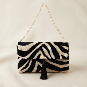 Puede incluir: Un bolso de mano rectangular con un patr&oacute;n de rayas de cebra en negro y crema y un acabado suave y texturizado. El bolso incluye una correa de cadena de metal dorado y una borla negra centrada en la solapa de cierre frontal.