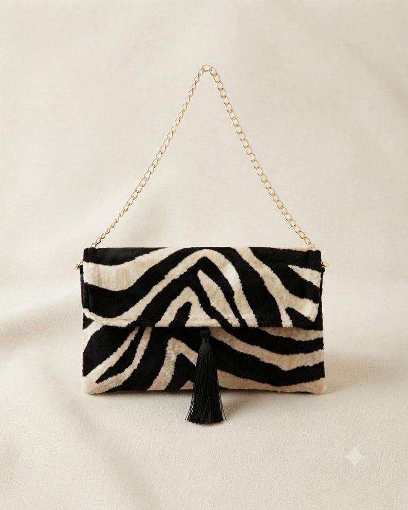 Puede incluir: Un bolso de mano rectangular con un patr&oacute;n de rayas de cebra en negro y crema y un acabado suave y texturizado. El bolso incluye una correa de cadena de metal dorado y una borla negra centrada en la solapa de cierre frontal.