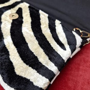 Puede incluir: Un bolso de mano con un patr&oacute;n de rayas de cebra en blanco y negro y un acabado texturizado similar al terciopelo. El bolso tiene una solapa con cierre magn&eacute;tico dorado y un forro interior negro con herrajes de color dorado.