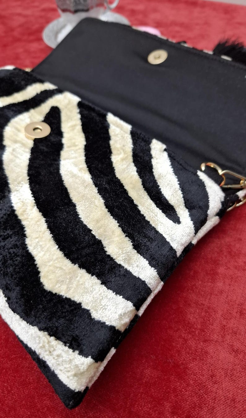 Puede incluir: Un bolso de mano con un patr&oacute;n de rayas de cebra en blanco y negro y un acabado texturizado similar al terciopelo. El bolso tiene una solapa con cierre magn&eacute;tico dorado y un forro interior negro con herrajes de color dorado.