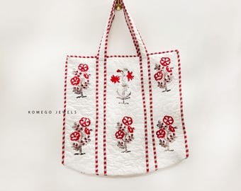 May include: A white quilted tote bag featuring a repeating red and grey floral block print pattern. The bag has two vertical red and white striped fabric straps and matching striped trim along the edges. The text KOMEGO JEWELS is printed in black.