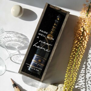 May include: A wooden box with a clear window, containing a bottle of Mo&euml;t & Chandon champagne. The box has a white inscription that reads "Pairs well with a new home".
