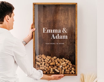 May include: A wooden frame with a glass front, displaying a collection of wine corks. The frame has a dark brown wood finish and the text "Emma & Adam Partners in Wine" is printed in white on the front.