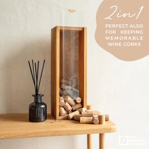 May include: A wooden wine cork holder with a clear acrylic top. The holder is filled with wine corks and is sitting on a wooden table. The text "2 in 1 PERFECT ALSO FOR KEEPING MEMORABLE WINE CORKS" is visible in the image.