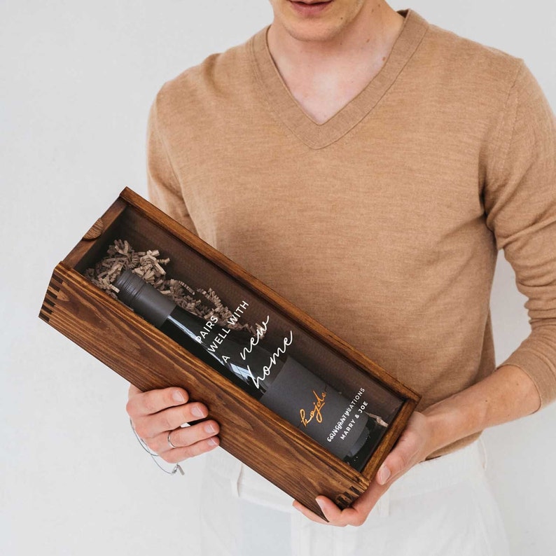 May include: A wooden box with a clear window, containing a bottle of wine with a label that reads "Pairs well with a new home".