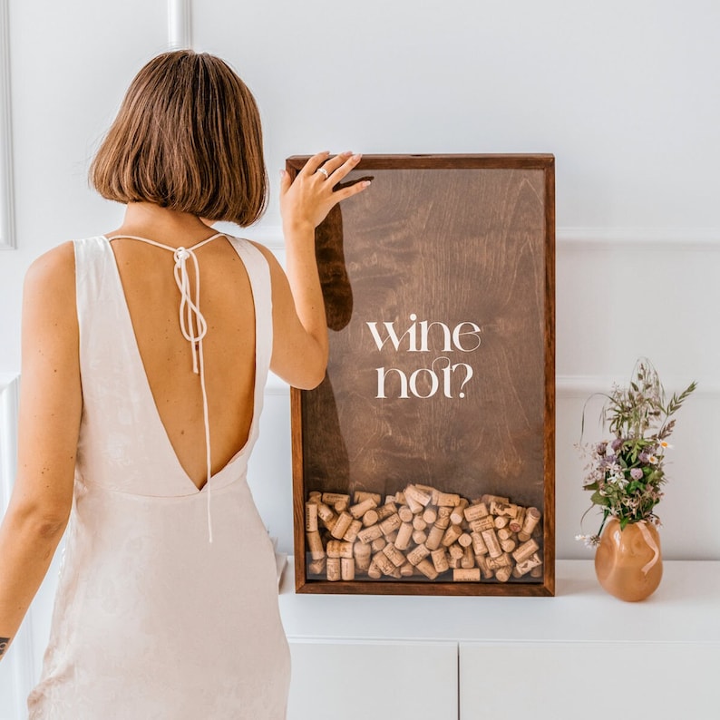May include: A wooden box with a glass front filled with wine corks. The box has the text "wine not?" printed on the front. A person is standing behind the box, wearing a white dress.