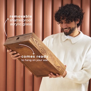 May include: A person holds a wooden frame with a removable shatterproof acrylic glass panel. The frame is ready to hang on a wall.