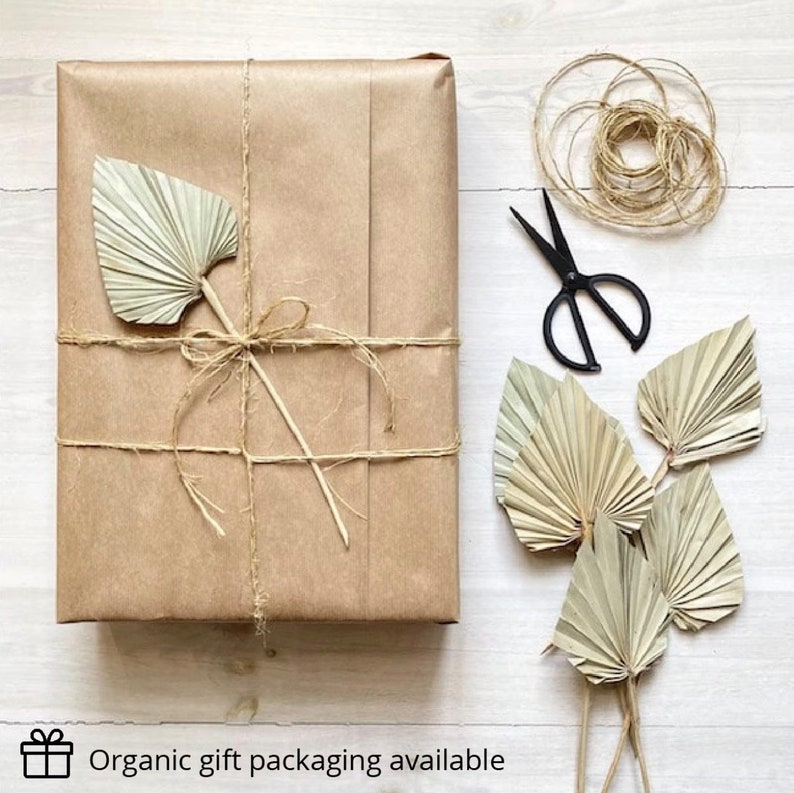May include: A brown paper package tied with twine and decorated with a dried palm leaf. The package is on a white wooden surface with a spool of twine, scissors, and more dried palm leaves.