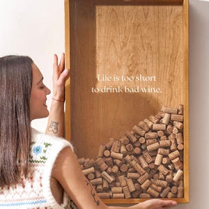 May include: A wooden shadow box with a glass front filled with wine corks. The shadow box has a white inscription that reads "Life is too short to drink bad wine."