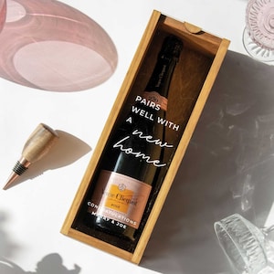 May include: A wooden box with a clear glass window, containing a bottle of Veuve Clicquot Rose champagne. The box has a white label that reads "Pairs well with a new home" and "Congratulations Marry & Joe".