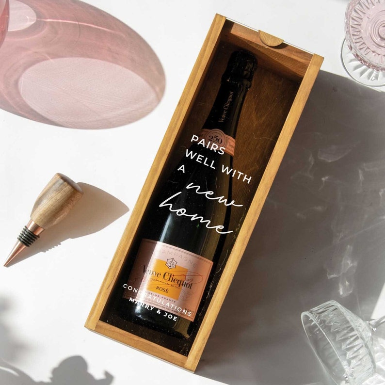 May include: A wooden box with a clear glass window, containing a bottle of Veuve Clicquot Rose champagne. The box has a white label that reads "Pairs well with a new home" and "Congratulations Marry & Joe".