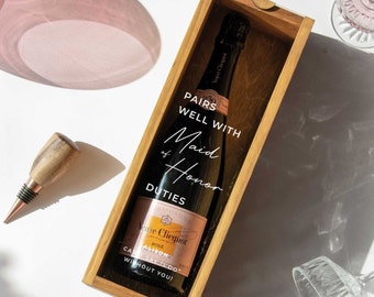 May include: A wooden box with a bottle of champagne inside. The box has a label that says "Pairs well with Maid of Honor Duties".