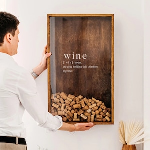 May include: A wooden wine cork holder with a glass front. The holder is filled with wine corks. The text "wine [ˈwaɪn] noun. the glue holding this shitshow together." is printed on the front of the holder.