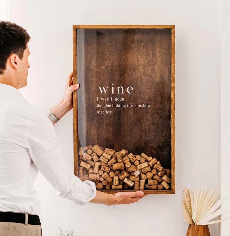May include: A wooden wine cork holder with a glass front. The holder is filled with wine corks. The text "wine [ˈwaɪn] noun. the glue holding this shitshow together." is printed on the front of the holder.