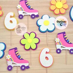 May include: A collection of colorful birthday cake toppers. The toppers include roller skates, flowers, and the number 6. The roller skates are pink with white accents and rainbow details. The flowers are yellow, pink, and blue.